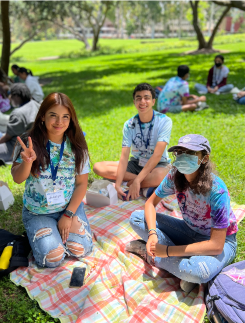 3 students sit on a blanket in Aldrich park. One holds up a peace sign
